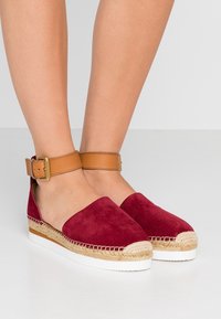 Espadrille en suède rouge avec une bride de cheville en cuir beige, dotée d'un semelle intermédiaire en jute tissé et d'une semelle extérieure en caoutchouc blanc. Texture lisse et design minimaliste.