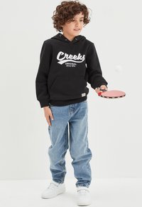 Felpa nera in cotone con logo bianco "Creeks", jeans in denim blu, tiene in mano una racchetta da ping pong rossa, indossa sneakers bianche, capelli ricci.
