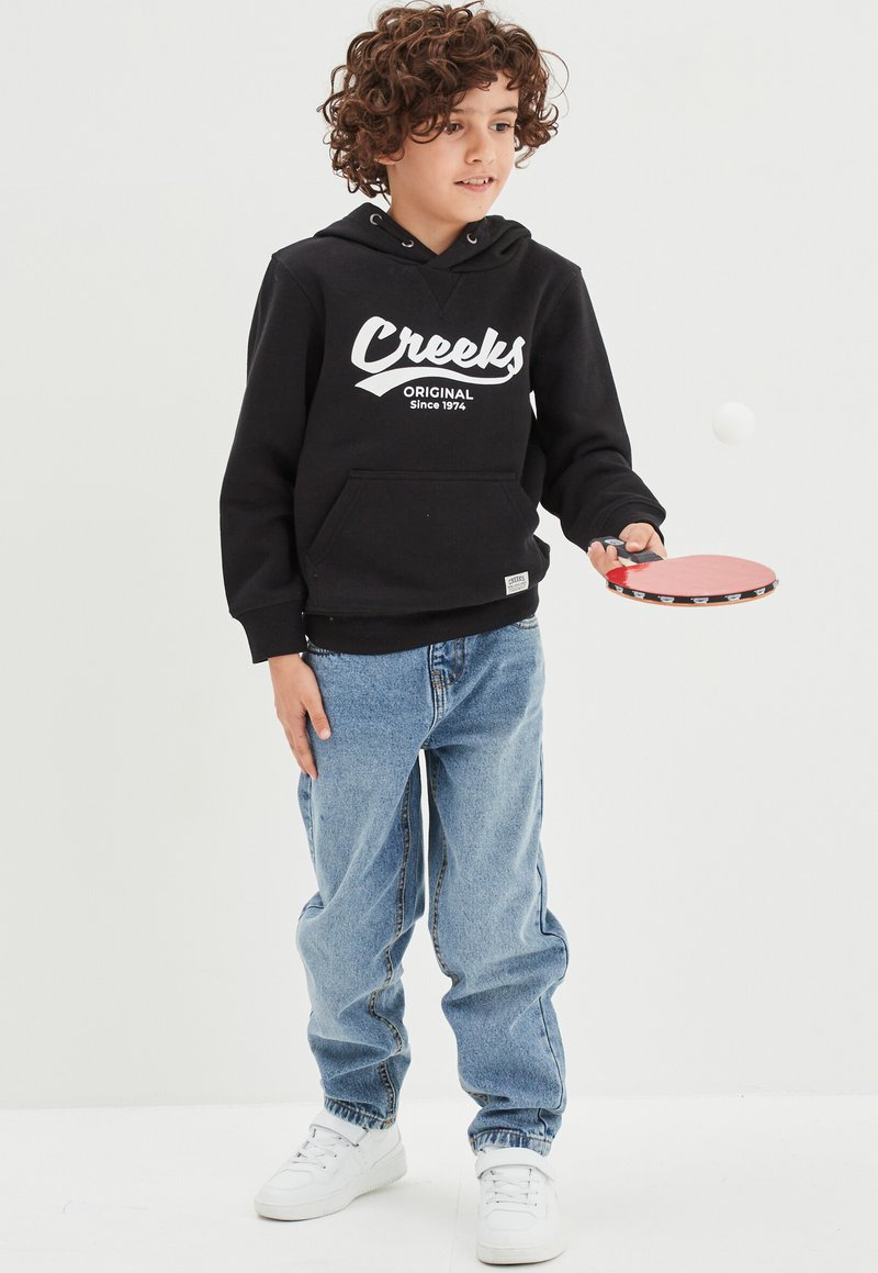 Felpa nera in cotone con logo bianco "Creeks", jeans in denim blu, tiene in mano una racchetta da ping pong rossa, indossa sneakers bianche, capelli ricci.