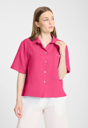 Vrouw draagt een felroze, korte mouwen blouse met knopen en witte broek, kijkt opzij tegen een effen achtergrond.