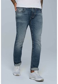Jeans de mezclilla azul con corte ajustado, ligero desgaste y costuras en contraste. Los dobladillos enrollados revelan unas zapatillas blancas con detalles de malla.