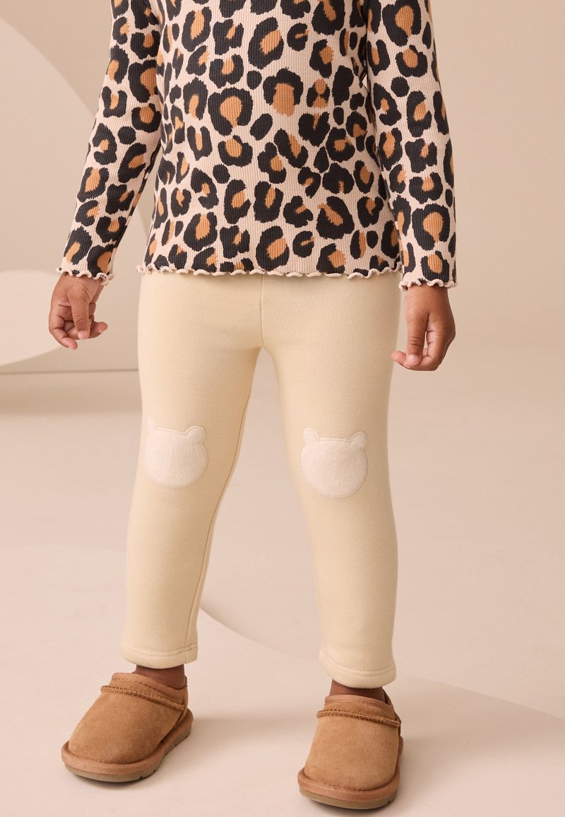 Enfant portant un haut à manches longues imprimé léopard, des leggings crème avec des patchs ours aux genoux et des chaussures marron à enfiler, debout sur un sol beige.