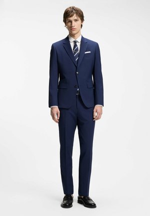 Giovane uomo che indossa un abito blu navy con camicia bianca, cravatta a righe e scarpe di pelle nere, in piedi davanti a uno sfondo bianco.