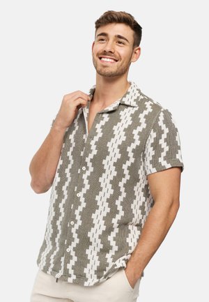 Homme souriant portant une chemise à manches courtes boutonnée avec des motifs géométriques gris et blancs, une main ajustant le col, l'autre dans la poche.