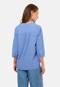 Blusa leggera azzurro chiaro a maniche lunghe con vestibilità rilassata, scollatura rotonda e dettaglio arricciato sul retro; abbinata a pantaloni in denim chiaro.