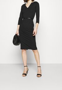 Robe noire, ajustée, de longueur genou avec un décolleté en V et des manches trois-quarts, accentuée par une étroite ceinture noire. Portée avec des sandales noires à talons.