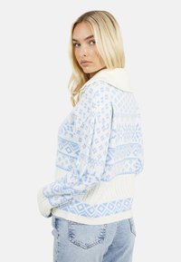 Gebreid wit trui met lichtblauwe geometrische patronen. Voorzien van een sjawlboord en geribbelde manchetten. Draagt met lichtblauwe jeans.