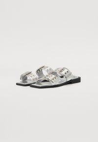 Sandales plates en argent en cuir métallisé avec deux larges sangles ornées de quincaillerie décorative et de rivets, montées sur une semelle noire.