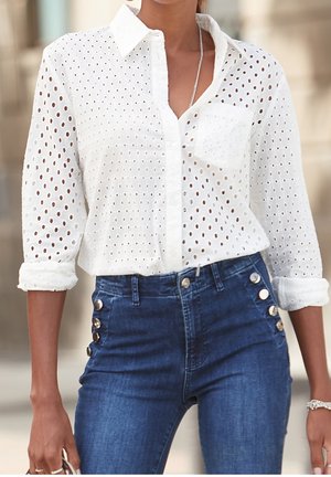 Hvid skjorte med knapper foran og åbent hulmønster, med lange ærmer som er rullet op. Kombineret med højtaljede, mørkeblå cowboybukser med knapper i siden.