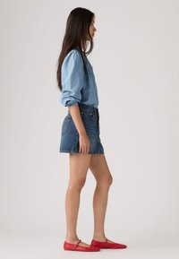 Ανοιχτό μπλε πουκάμισο από τζιν με μακριά μανίκια, σορτς από μπλε τζιν με ψηλή μέση, κόκκινα δερμάτινα μπαλαρίνες με λουράκι, όρθια πλευρική όψη.