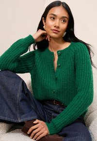 Zelený pletený kardigan s žebrovanou texturou, předním zapínáním na knoflíky a dlouhými rukávy. V kombinaci s tmavými džíny, sedící na světlém povrchu.