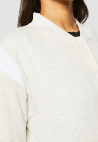 Femme portant une veste beige clair boutonnée avec un col montant et une bande diagonale blanche sur la manche, par-dessus un haut noir.