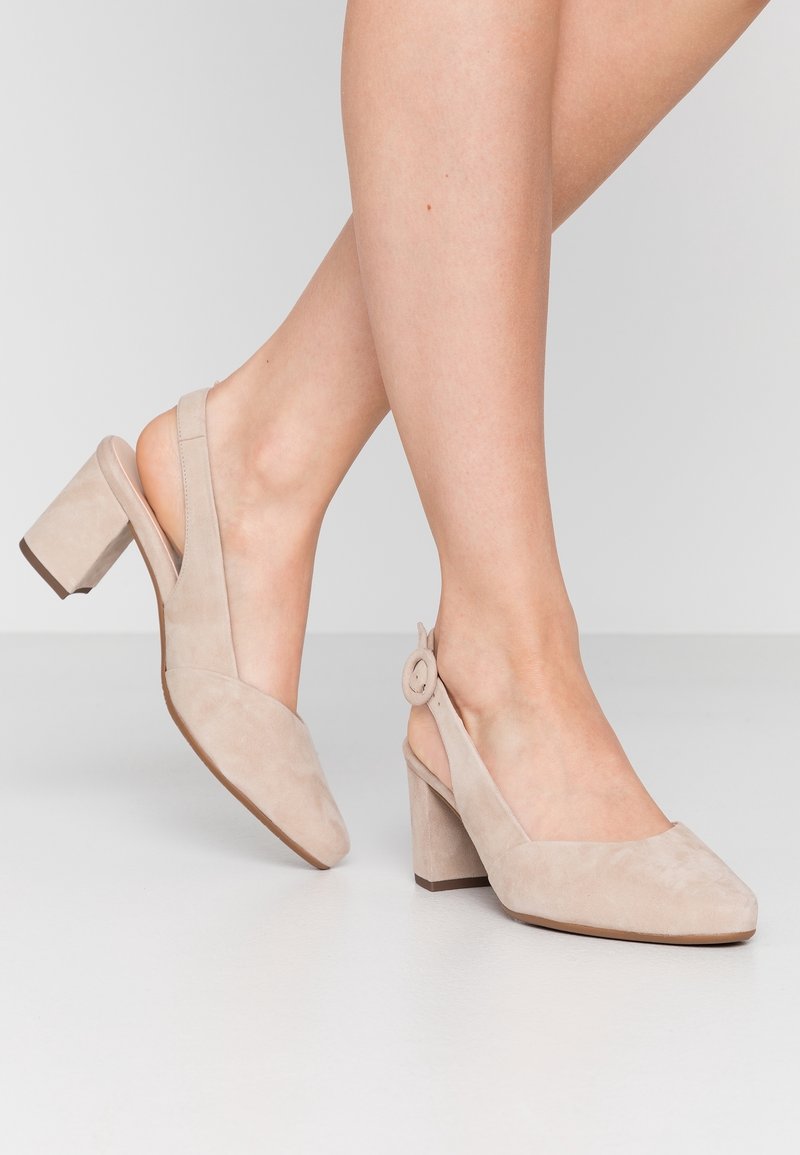 Talons slingbacks en suède beige avec un bout carré, un talon épais et un accent circulaire sur la sangle ajustable. Texture lisse, design minimal.