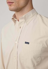 Beige knoopoverhemd van katoen, met een kraag, een borstzak met een marineblauw label en knopen in een bijpassende kleur.