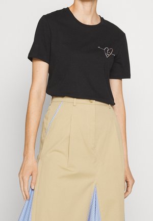 Person trägt ein schwarzes T-Shirt mit einer kleinen lächelnden Herzgrafik in der Nähe der Brust und einen beigen Rock mit blauen gestreiften Falten.