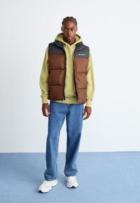 Puffervest met een bruine romp en zwarte schouders, gedragen over een lichtgroene hoodie, gecombineerd met blauwe jeans en witte sneakers.