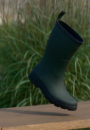 Botte en caoutchouc vert foncé suspendue au-dessus d'une surface en bois avec de hautes herbes vertes en arrière-plan.