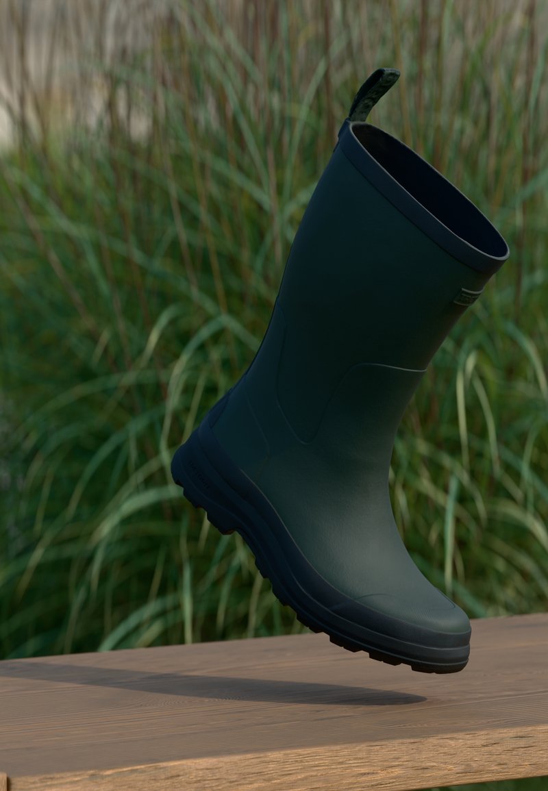 Donkergroene rubberen laars zweeft boven een houten oppervlak met hoge groene gras op de achtergrond.