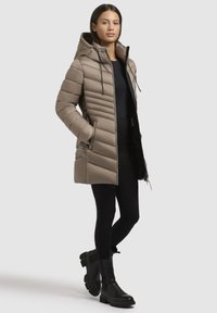 Pufferjack in taupe met een doorgestikt ontwerp, verstelbare capuchon en zijzakken. Gecombineerd met zwarteleggingen en chunky zwarte enkellaarsjes.