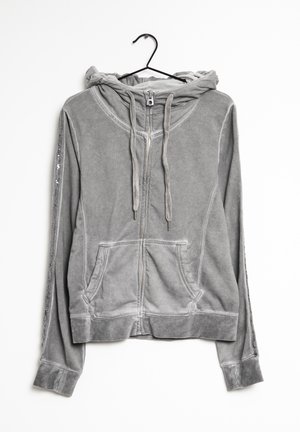 Harmaa vetoketjullinen huppari, jossa nyörit ja etutaskut, ripustettuna mustalle henkarille valkoista taustaa vasten.