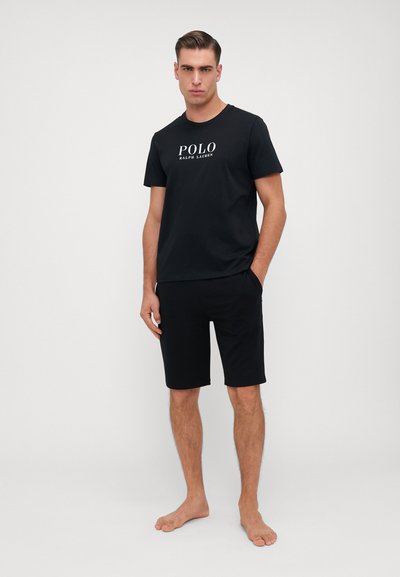 Cămașă albă din bumbac cu imprimeu "POLO RALPH LAUREN" în alb, combinată cu pantaloni scurți negri, evidențiind o siluetă relaxată și casuală.
