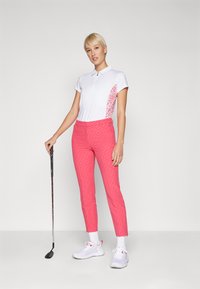 Balts polo krekls ar rozā rakstainu sānu, apvienots ar rozā rakstainām biksēm. Modelis tur golfa nūju un valkā baltas sporta kurpes.