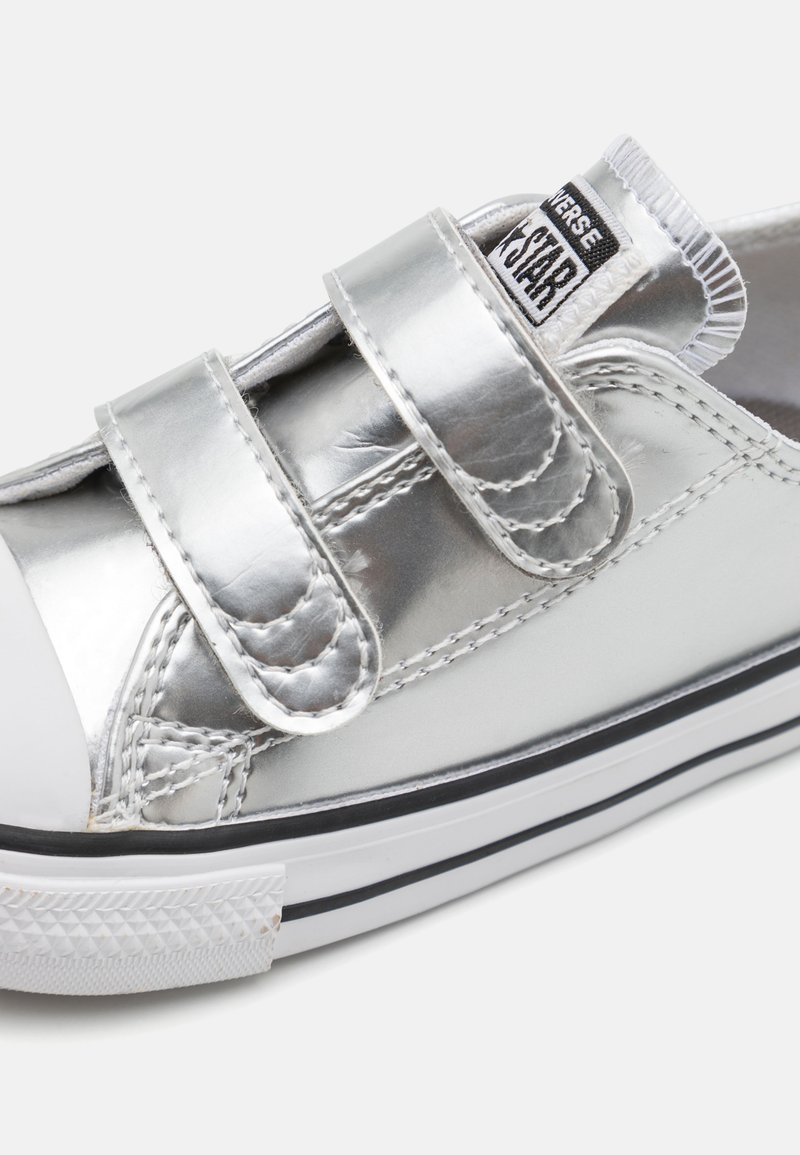 Zilveren metallic sneaker met dubbele klittenbandsluitingen, witte rubberen neuskap en zwart-wit gestreepte zool. Glad textuur en gestikte accenten.