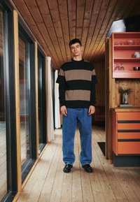 Pull rayé noir et marron, associé à un jean bleu ample, debout sur un sol en bois dans un intérieur moderne avec des armoires orange.