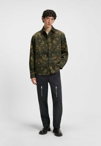 Grön mönstrad skjorta med ett botaniskt mönster och guldiga accenter, parat med mörka byxor med dragkedjefickor och en slät textur.
