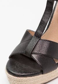 Sandale compensée noire avec des brides texturées croisées sur les orteils et une bride à la cheville, avec une semelle plateforme tissée en fibre naturelle.