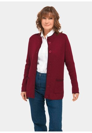 Bordeaux kleurige cardigan met knopen, ronde halslijn, twee voorzakken en contrasterende stiksels, gecombineerd met een wit shirt en blauwe spijkerbroek.