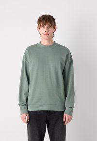 Sudadera verde con cuello redondo y mangas largas, con un logo sutil en el pecho. Combinada con jeans oscuros.