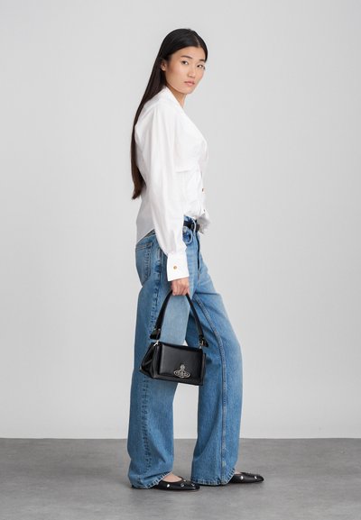 Fekete kézi táska strukturált formával, lapos felsőfogantyúval és logó részletekkel. A modellen fehér ing és világoskék, bő szárú farmernadrág van. Lapos fekete cipő.