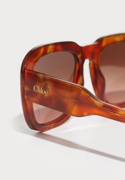 Lunettes de soleil en écaille avec une monture épaisse et rectangulaire, verres dégradés marron, et logo gravé en or sur la branche, texture lisse.