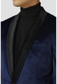 Blazer in velluto blu navy con colletto a scialle in satin nero, design a petto singolo e tasca sinistra discreta. La texture interna è liscia.