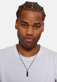 Zwarte rechthoekige hangerketting op een dunne zwarte ketting, gedragen over een wit T-shirt. Gladde metalen afwerking, minimalistisch ontwerp.