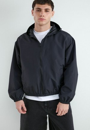 Ung mand iført en sort lynlåshættetrøje over en hvid t-shirt og sorte bukser, stående mod en ensartet lys baggrund.