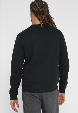 Mann mit langen Dreadlocks, die nach hinten gebunden sind, trägt ein schlichtes schwarzes Sweatshirt und graue Hose, steht mit dem Rücken zur Kamera.