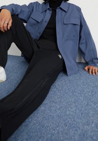 Blå skjorta med knappar och två bröstfickor, kombinerad med svarta byxor med sidoremsor och en logotyp, bärs med vita sneakers på en blå yta.