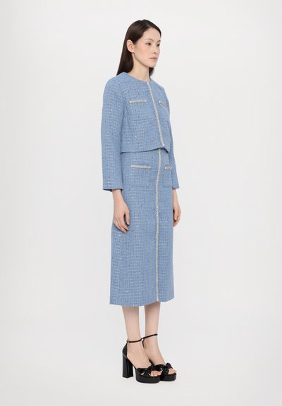 Blaues Tweed-Zweiteiler mit einer kurzen Jacke, silbernem Reißverschluss und Taschen. Kombiniert mit schwarzen Plateausandalen mit Schleifen.