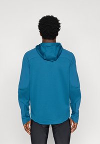 Hanorac sportiv albastru cu un model texturat și cu nervuri. Prezintă o glugă, mâneci lungi și manșete ajustabile. Un design general ajustat.