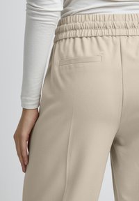 Beige nadrágok puha, texturált anyagból. Gumírozott derékkal és egy hátsó zsebbel rendelkezik. Oldalnézetből látható.