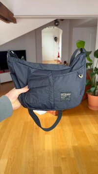 Mala de ombro preta em nylon com formato curvo, apresentando dois bolsos exteriores e uma etiqueta da marca. Textura suave e alça ajustável.