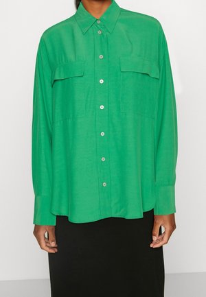 Personne portant une chemise boutonnée vert vif avec deux poches poitrine et manches longues, associée à une jupe noire.