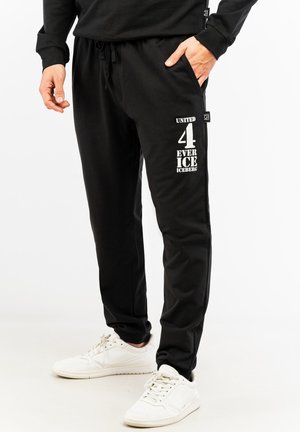 Schwarze Jogginghosen aus weichem Material mit elastischem Bund, seitlichen Taschen und bedrucktem Textdesign in Weiß am linken Oberschenkel.