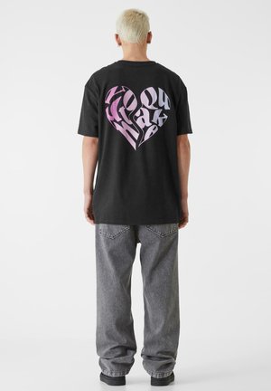 Sorte oversized t-skjorte med et rosa gradient hjertegrafikk og stilisert tekst på baksiden. Kombinert med grå baggy-jeans og svarte sko.