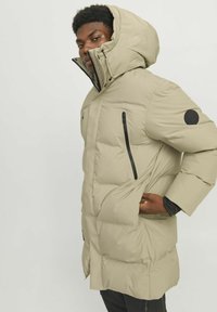 Beige padded parka with a high collar and hood, featuring black zippered side pockets and a round logo patch on the left sleeve.