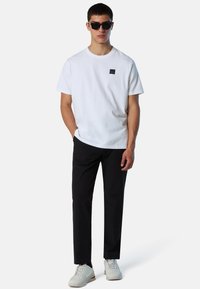 T-shirt bianca in cotone con un piccolo logo nero, abbinata a pantaloni neri e sneakers bianche testurizzate, con occhiali da sole.