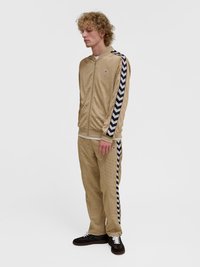 Mlada oseba z kodrastimi blond lasmi, oblečena v bež trenirko z mornarsko modrimi chevron črtami na rokavih in hlačah, stoji na belem ozadju.