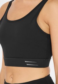 Musta urheiluliivi, joka on valmistettu sileästä ja joustavasta kankaasta. Siinä on pyöreä pääntie ja hillitty raidallinen kuvio helmassa.